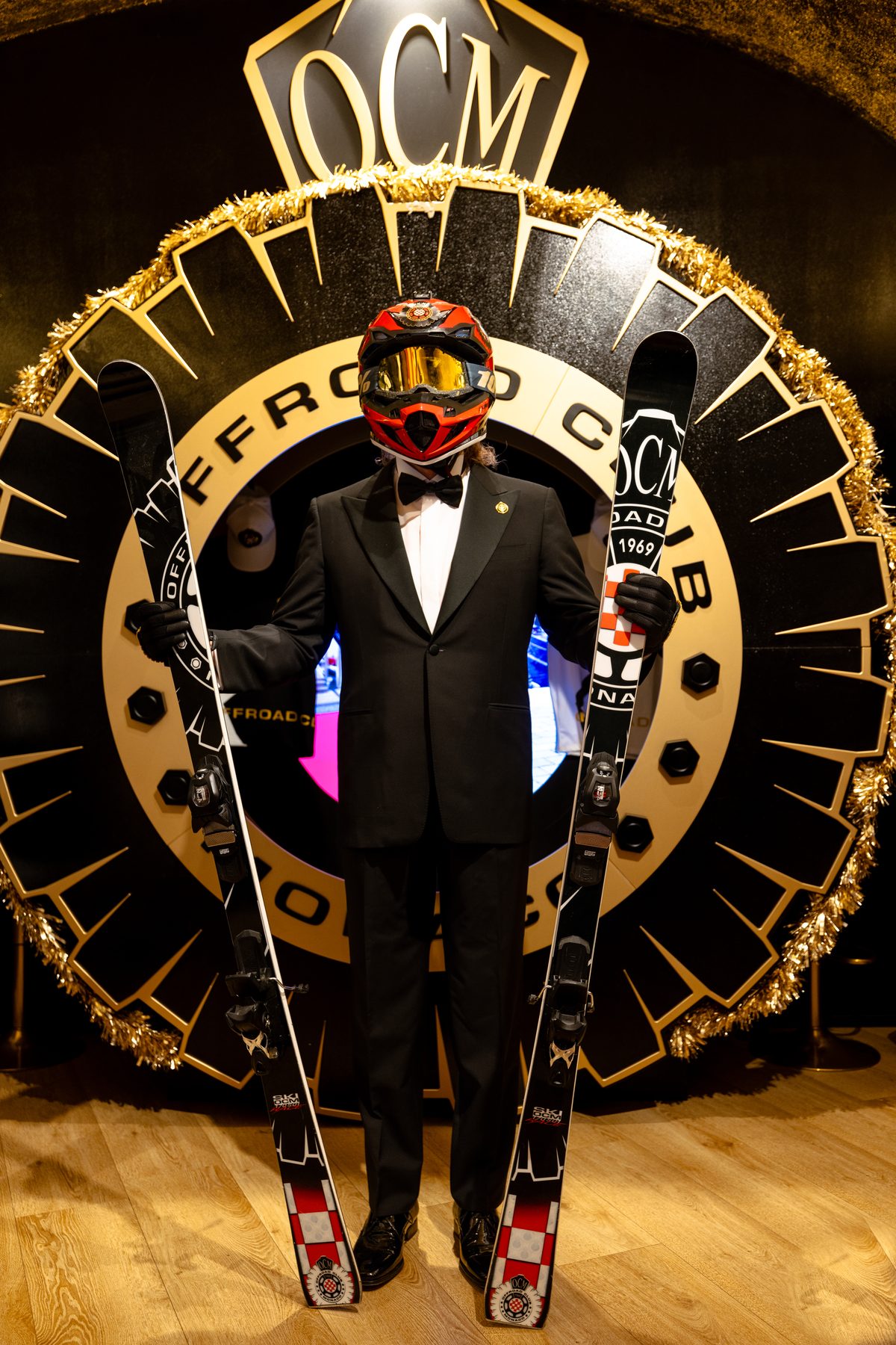 Mascotte Offroad Club Monaco in smoking con casco e sci davanti al grande logo OCM